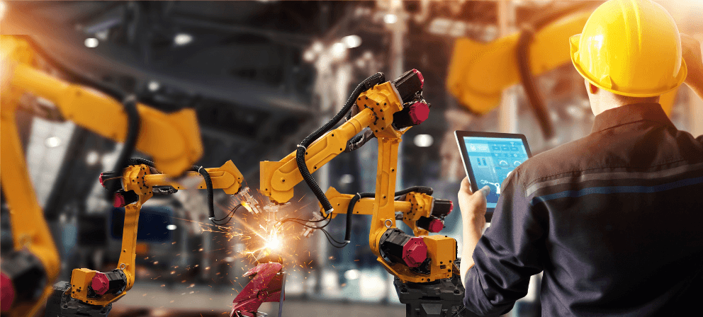 A technician in a yellow hard hat uses a tablet to control robotic arms welding in an industry 5.0 setting. The robotic arms, with yellow bodies and red and black components, emit visible sparks from welding. The background is filled with more machinery in a bustling factory environment.