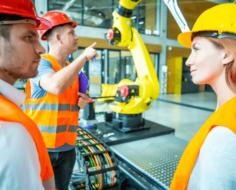 Three engineers wearing orange safety vests and yellow hard hats are in a factory setting, discussing a large yellow robotic arm. One person is pointing at it while others listen attentively, standing close to cable and machinery equipment, embodying the principles of Industry 5.0 collaboration.